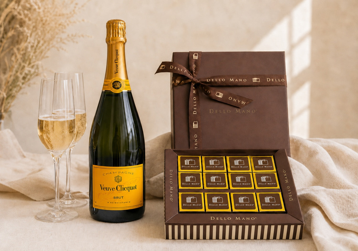 Veuve Clicquot Champagne with two filled flutes and a Dello Mano brownie gift box containing gold foil-wrapped brownies with Dello Mano labels, styled on soft neutral linen.
