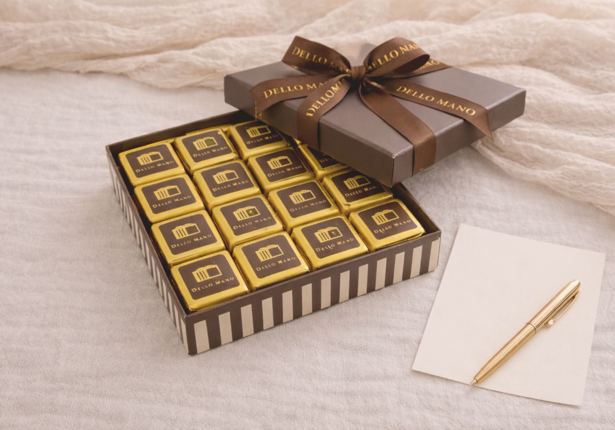 Open Dello Mano chocolate gift box with gold-wrapped brownies, brown grosgrain ribbon, and gift card on linen background