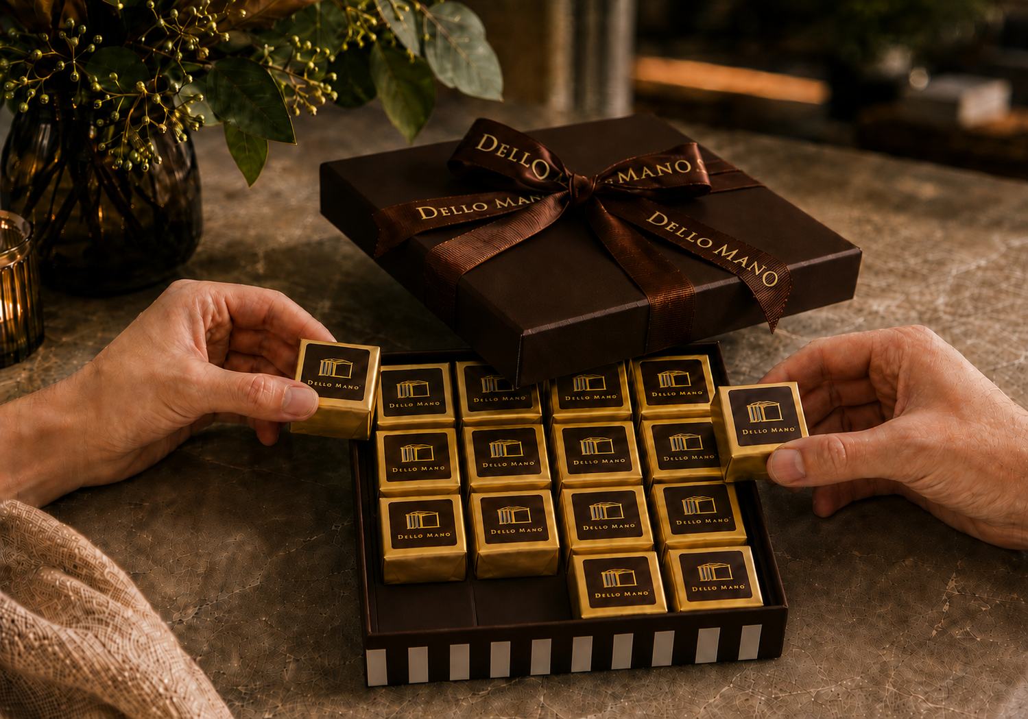 Hands selecting individually wrapped Dello Mano brownie cubes from an open chocolate gift box