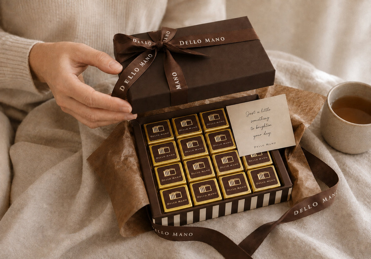 Dello Mano chocolate gift box being opened by hand, with gold-wrapped brownies, tissue paper and a personal message card on soft cream linen.