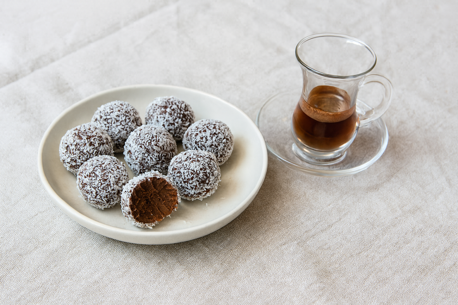 Handmade coconut-coated rum balls on a modern handmade plate with a Turkish-style glass of espresso, set on natural linen in cool white light, calm aesthetic.