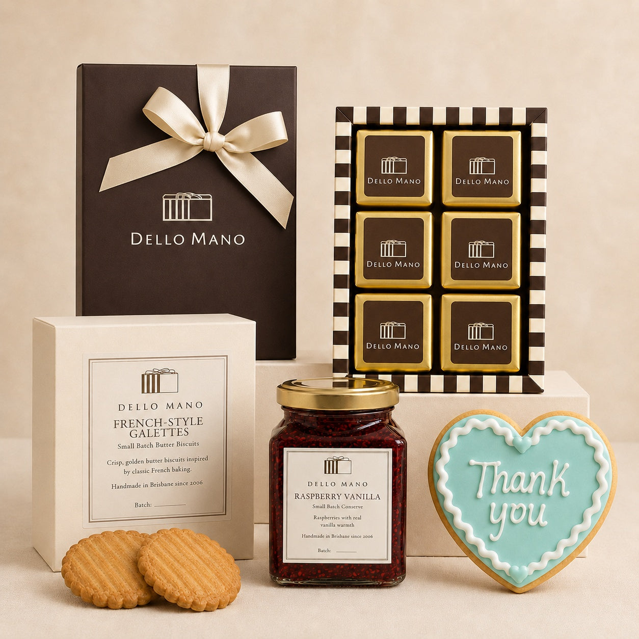 Dello Mano The Thank You Hamper featuring a 6-piece brownie gift box, French-style galettes, Raspberry Vanilla conserve and a pale blue iced thank you cookie, styled on neutral plinths.