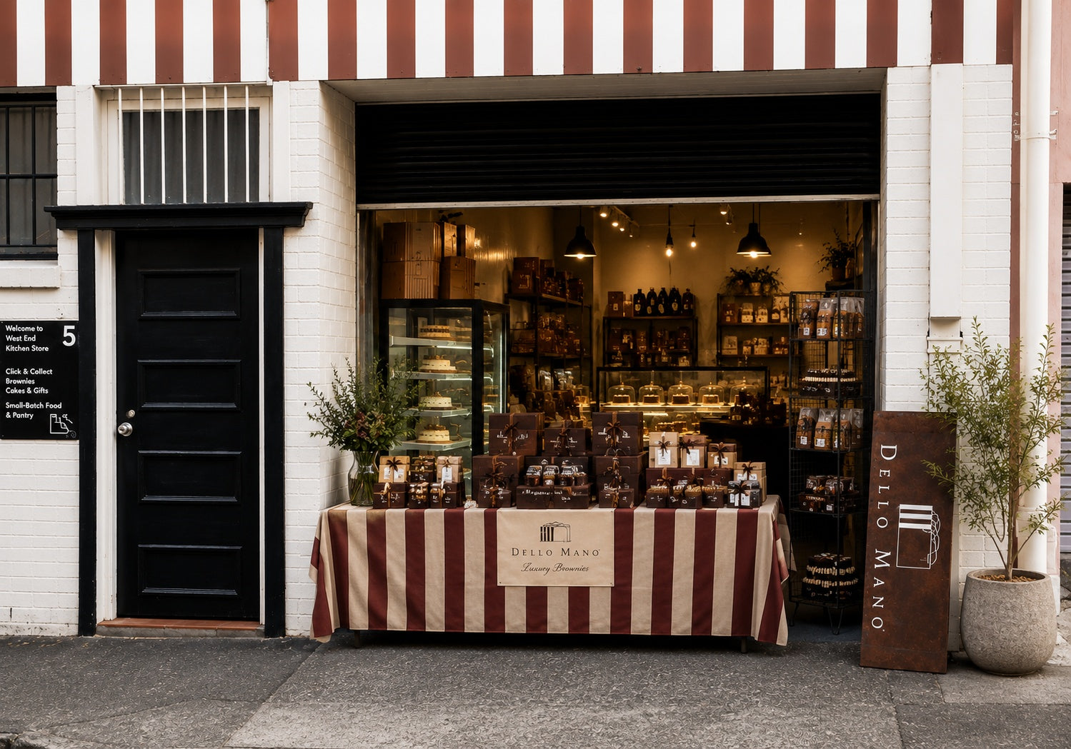Mock up of Dello Mano West End Kitchen Store with an open entrance, handmade gifts and cakes displayed on a striped table, and warm shelving visible inside.
