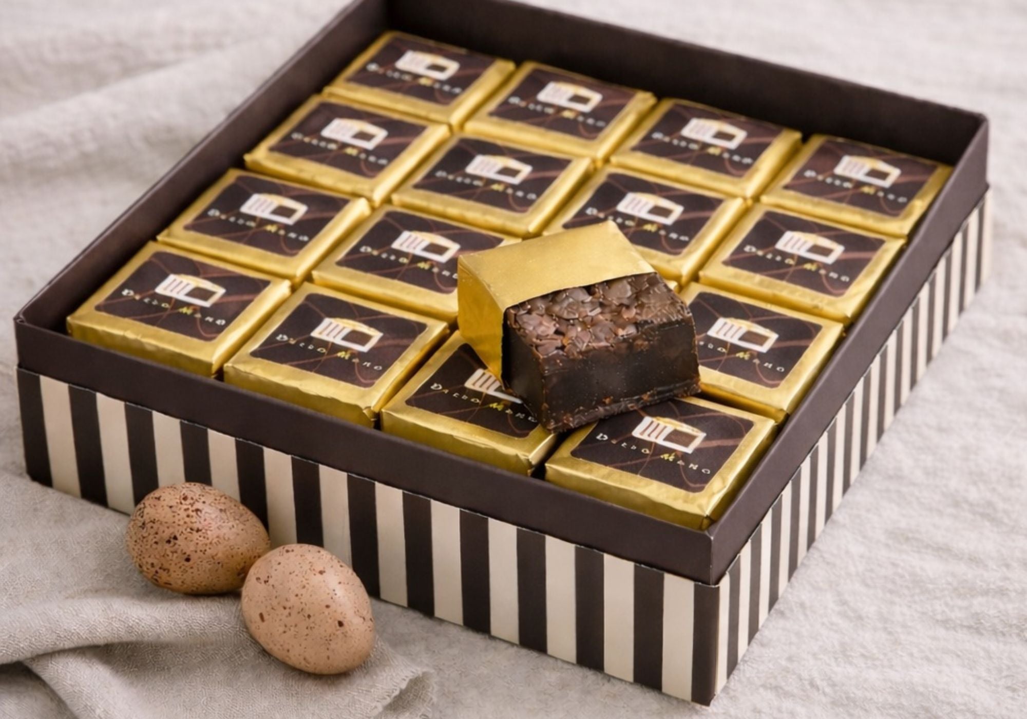 Dello Mano brownie gift box with gold foil wrapped chocolate brownies and speckled Easter eggs on natural linen background.
