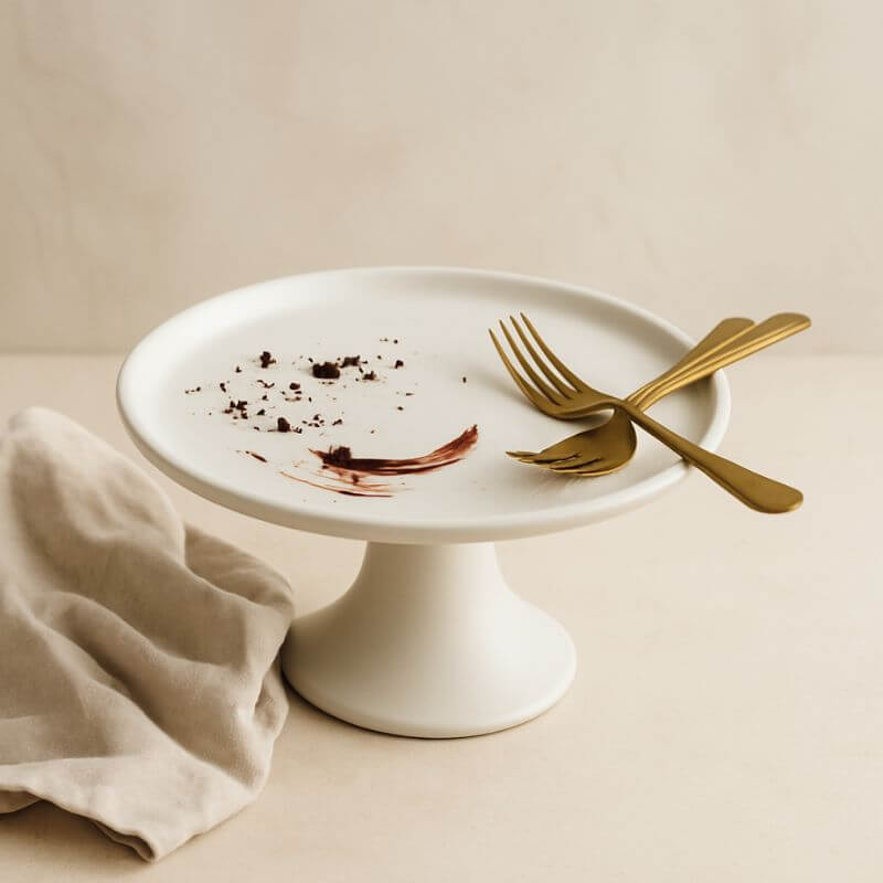 White ceramic cake stand with chocolate crumbs and used gold forks, suggesting an elegant dessert just enjoyed.