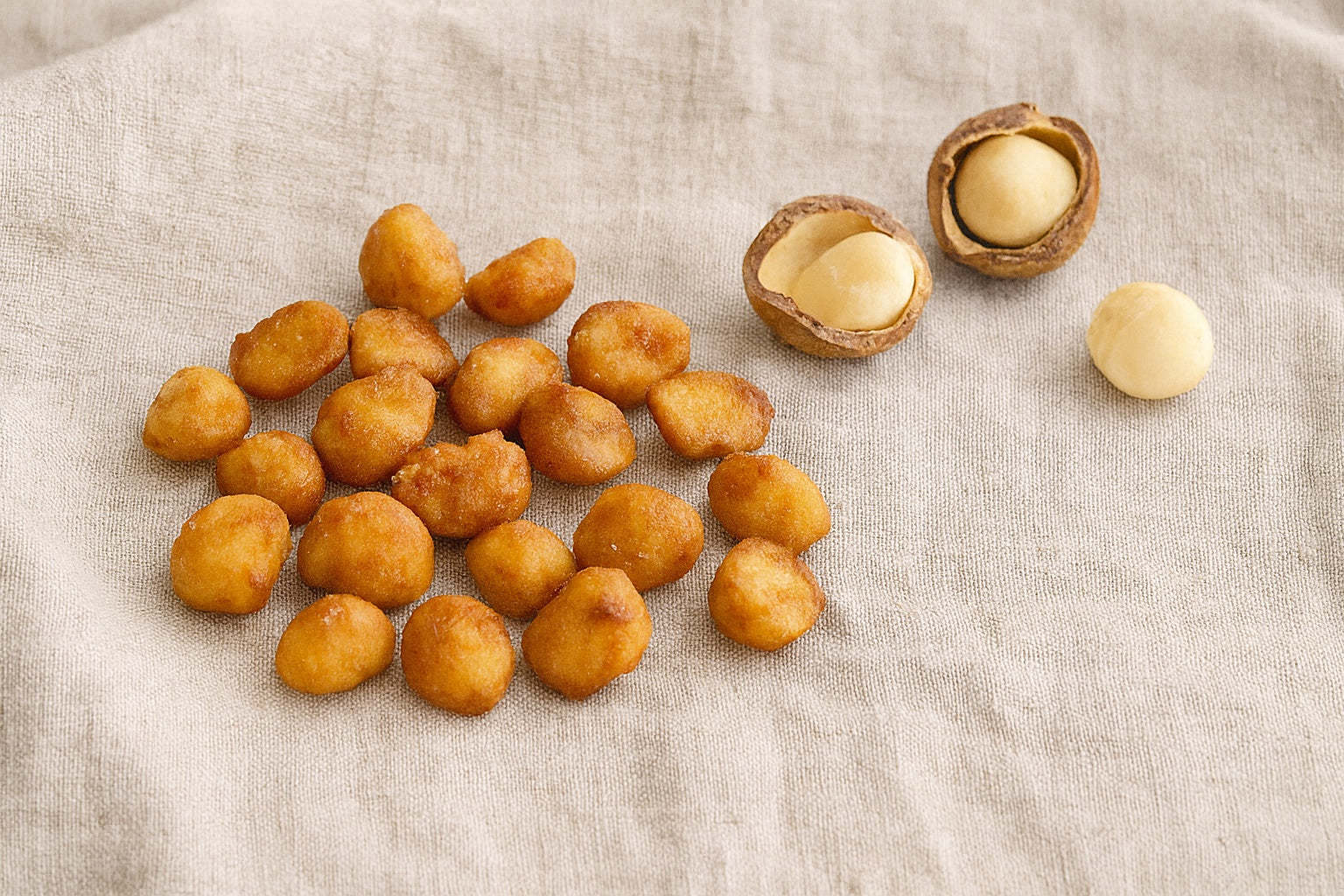 Handmade honey caramelised macadamias scattered on natural linen cloth, with cracked raw macadamia shells showing creamy kernels, highlighting golden caramel gloss