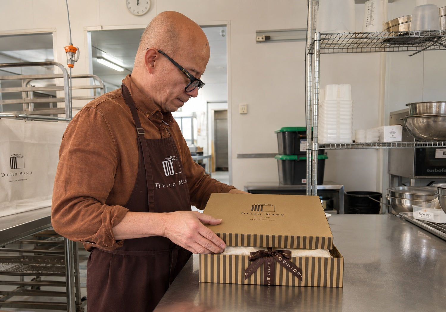 Bien closing a striped Dello Mano hamper box on a stainless-steel bench in the West End kitchen, with cream tissue and branded ribbon visible.