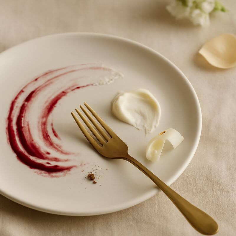 White plate with raspberry smear, gold fork, and small white flowers on a beige background