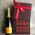 A bottle of Veuve Cliquot Champagne lying beside and open box of gold foiled brownies. The chocolate gift box lid has a brown ribbon that says Dello Mano