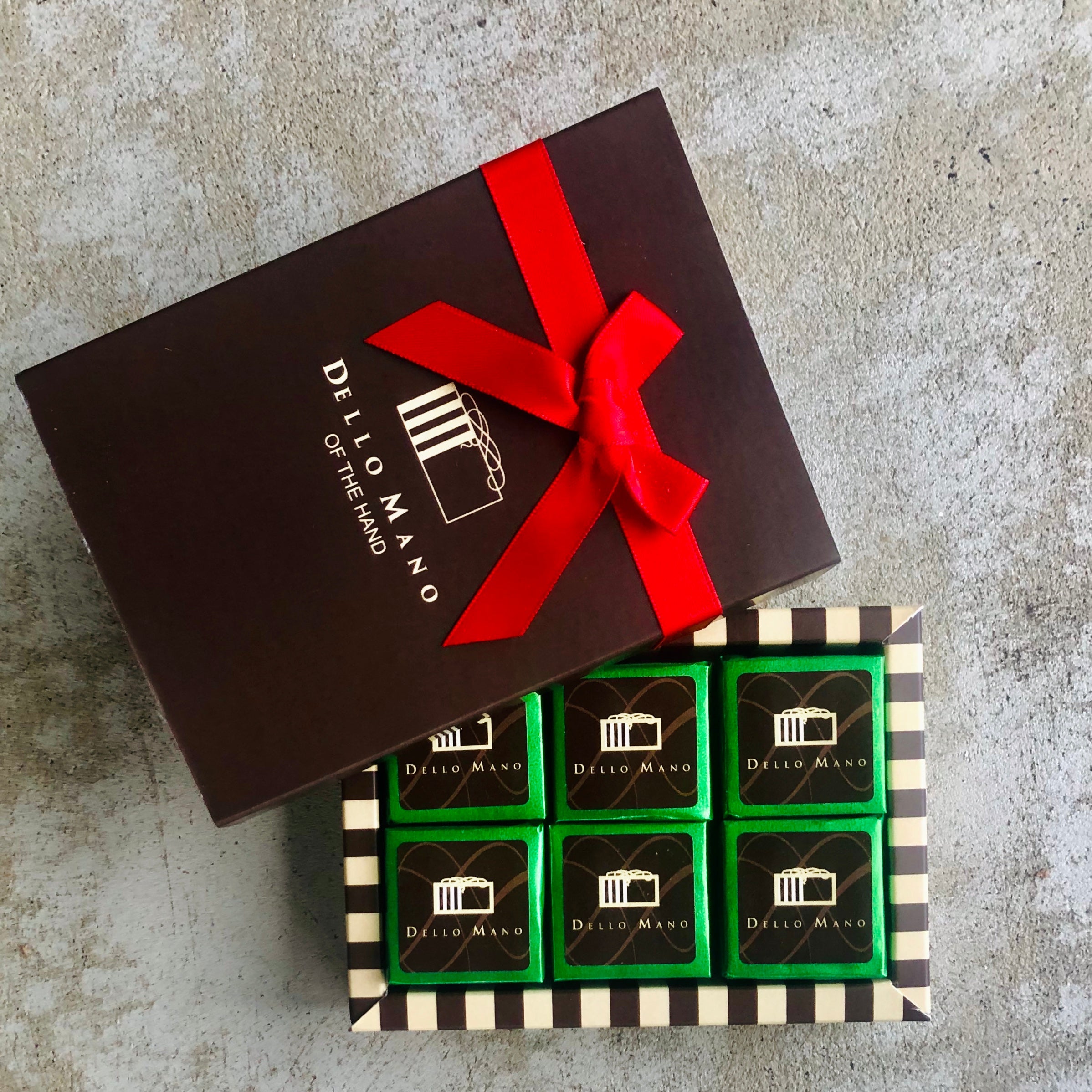 A small chocolate gift box of Vegan green foiled brownies. The lid has red ribbon and a logo and the words Dello Mano of the hand.