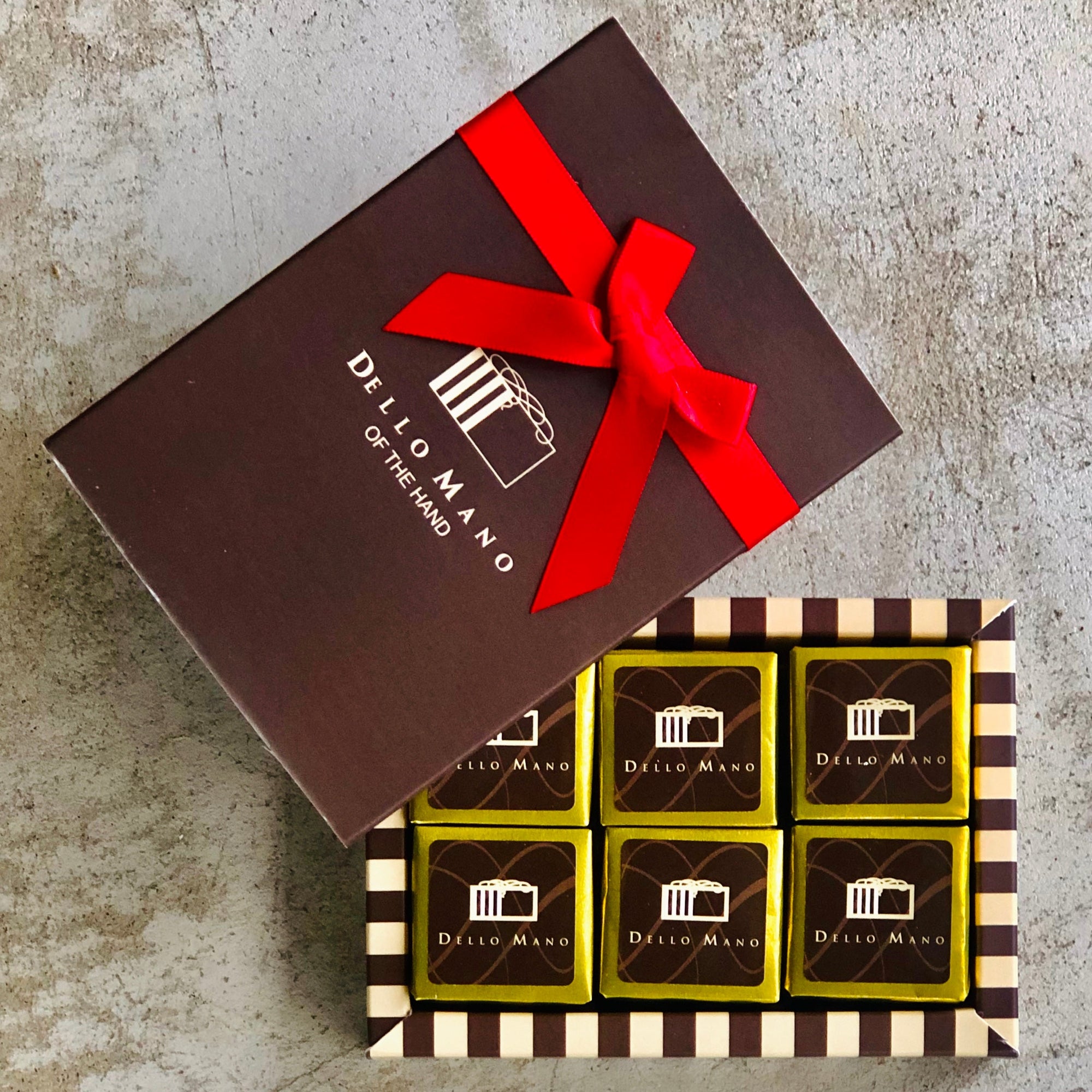 A small gift box of gold foiled brownies . A brown box lid lies at the side with a red ribbon and the words Dello Mano of the hand on the box.