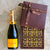 A bottle of Veuve Cliquot Champagne lying beside and open box of gold foiled brownies. The chocolate gift box lid has a brown ribbon that says Dello Mano