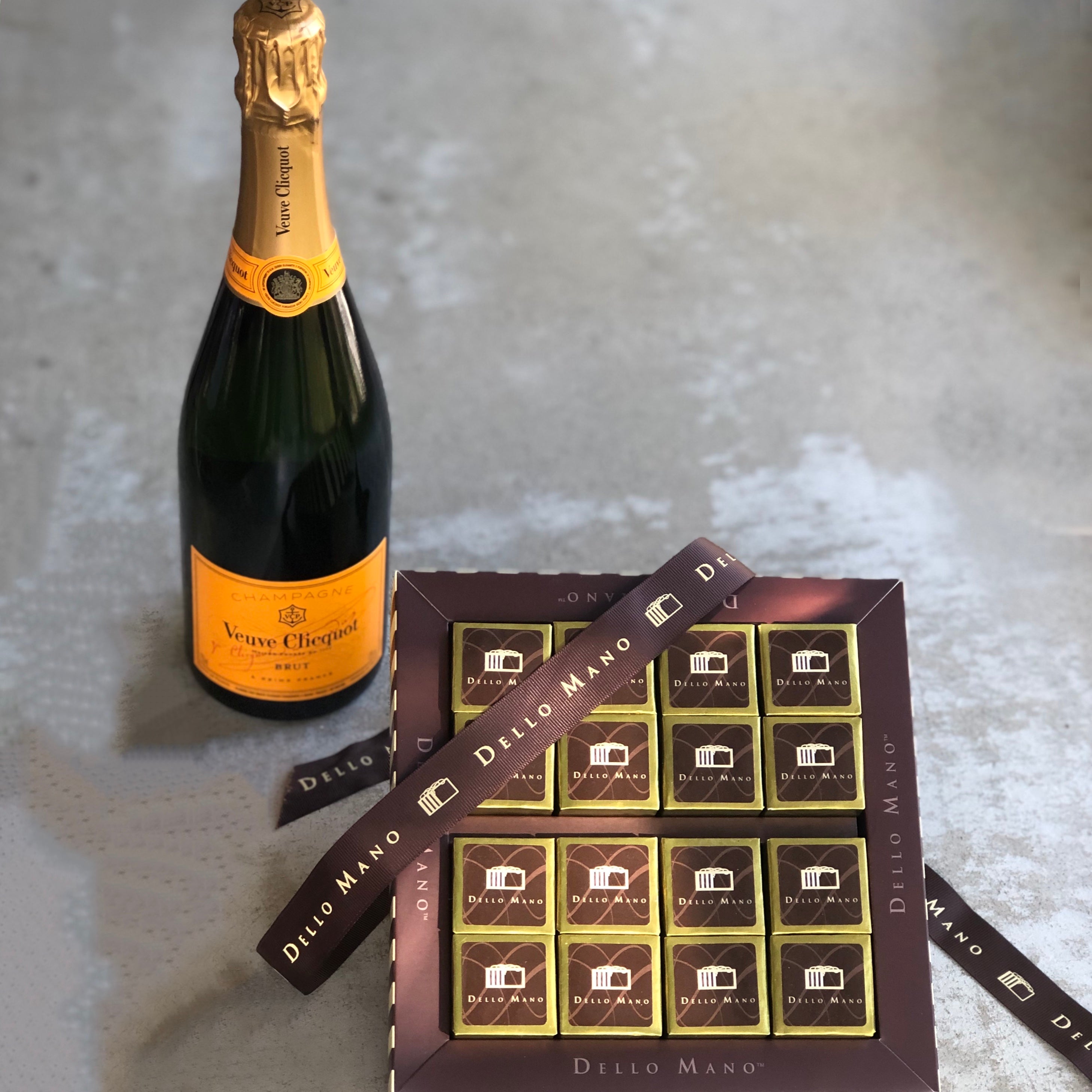 Bubbles (champagne) and brownies sitting on a concrete bench top. A ribbon with the words Dello Mano is draped across the top of the chocolate gift box