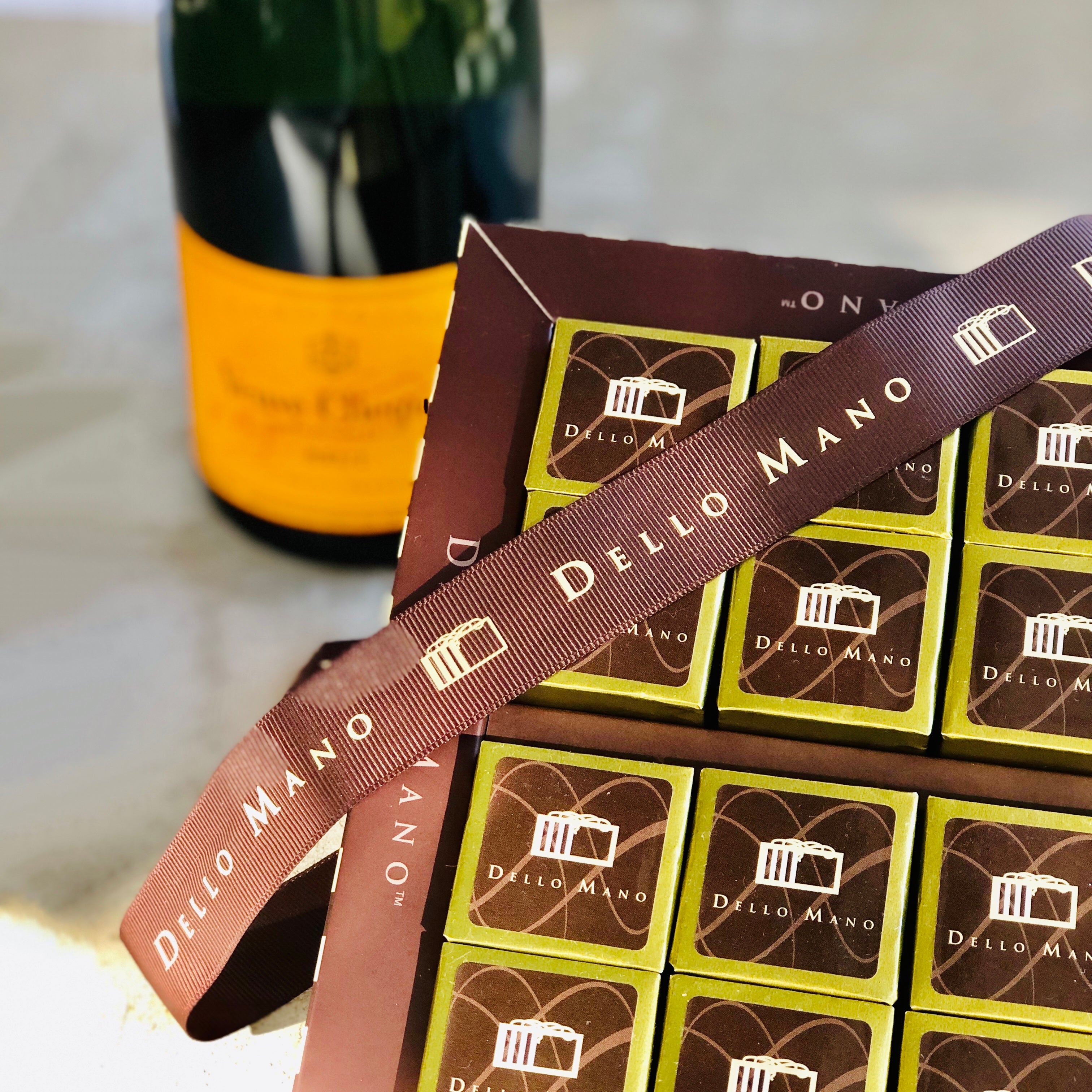 A champagne bottle with brownies. The Champagne bottle has an orange label and is blurred in the background . The gold foiled brownies have a brown ribbon across them and the ribbon says Dello Mano