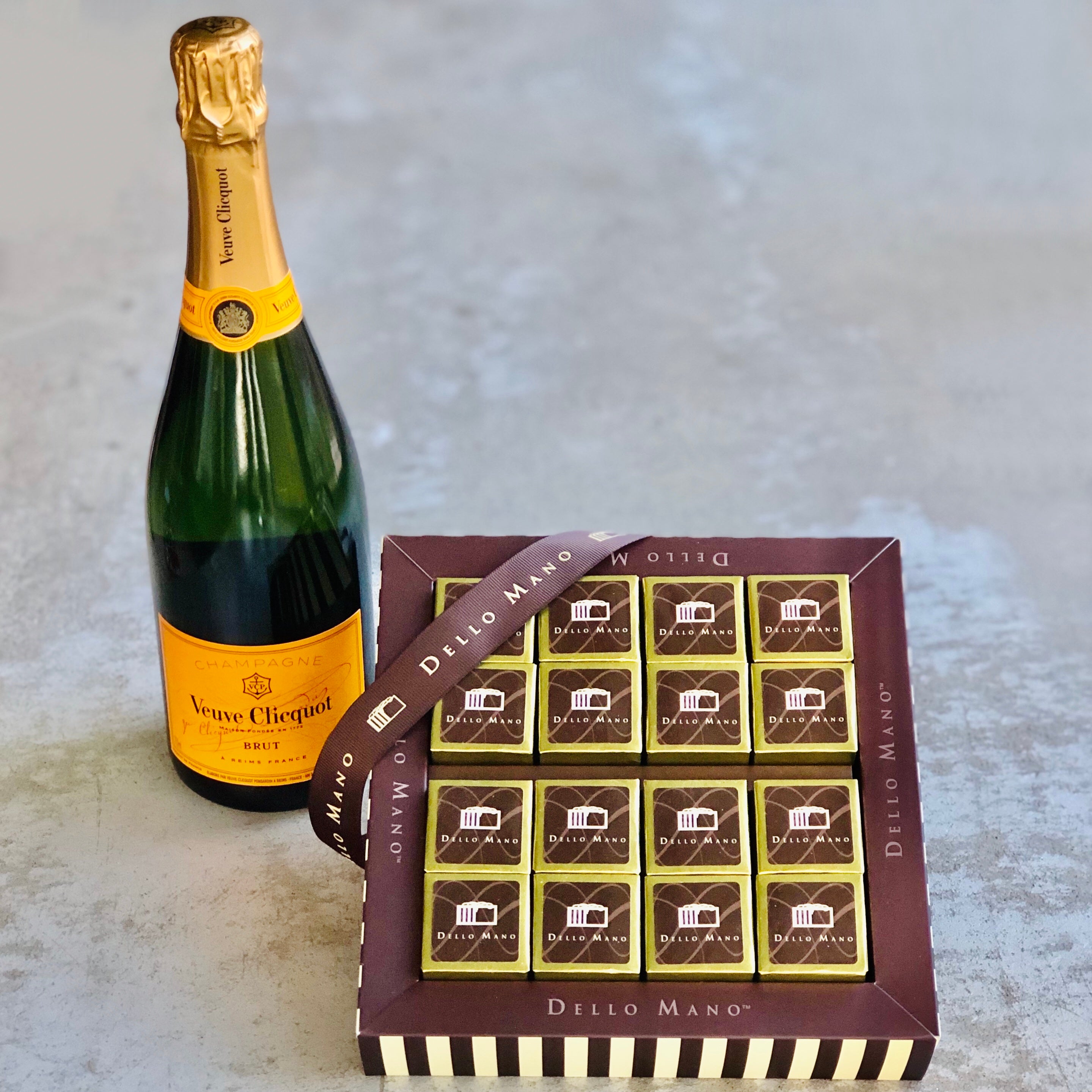 Bottle of bubbles and box of brownies. The champagne is standing up and has a label saying Veuve Cliquot. The gold foiled brownies are in a brown chocolate box with words saying Dello Mano