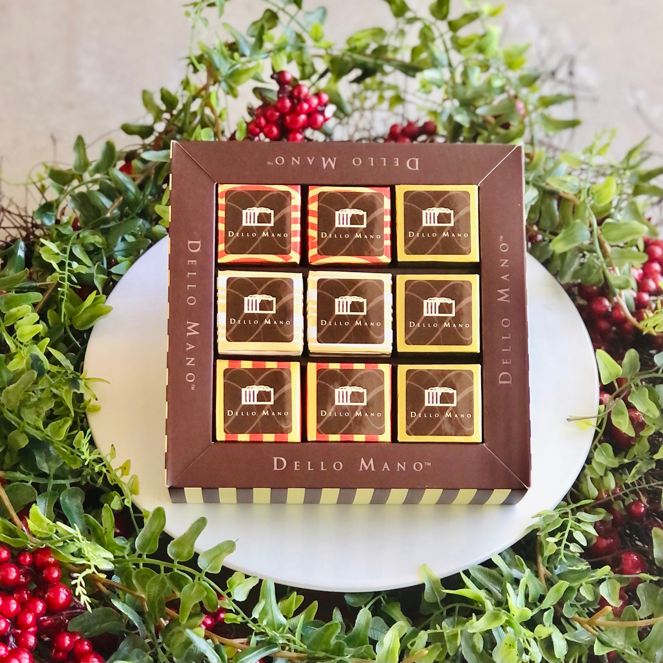 A Christmas Brownie sampler box of 9 brownies. The box says Dello Mano and is surrounded by greenery and red berries