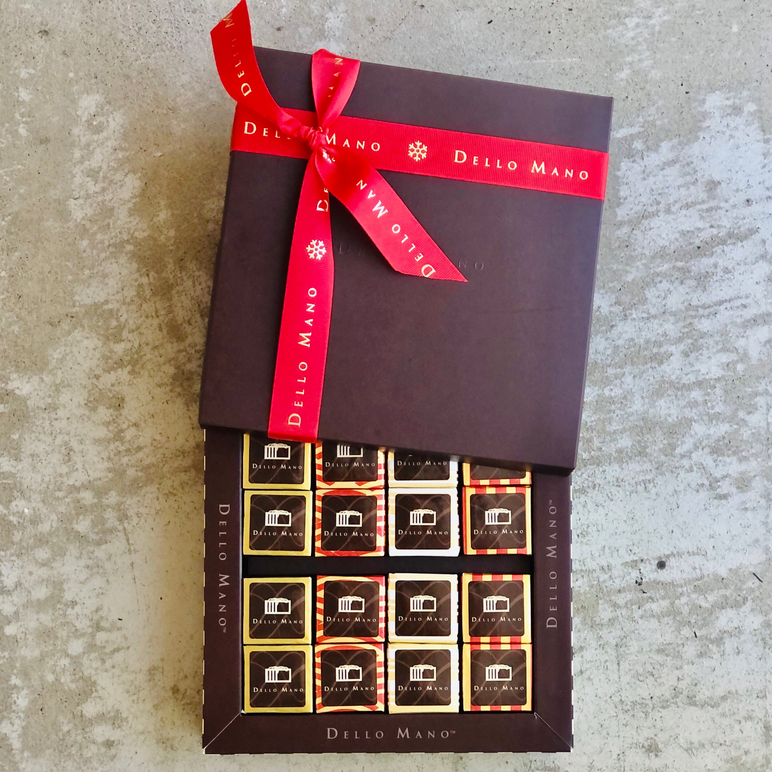 An open chocolate gift box filled with Christmas Brownies. Each individually wrapped in foil. The box lid sits on top open with a red Christmas ribbon that says Dello Mano