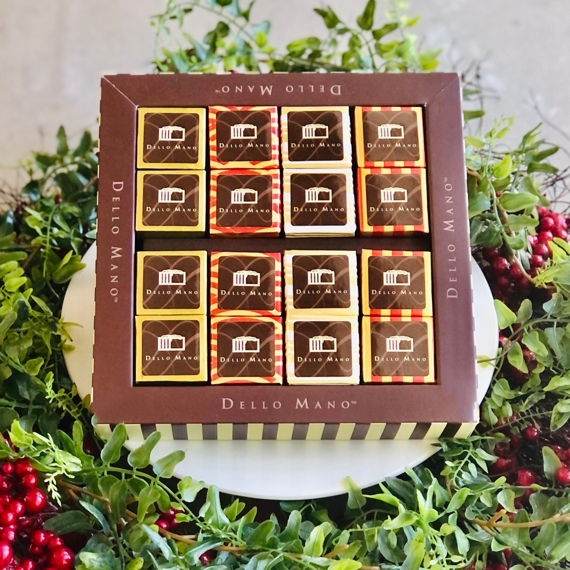 A Christmas Brownie sampler gift box filled with 16 foil covered brownies. The box says Dello Mano with words and is surrounded by greenery and red berries