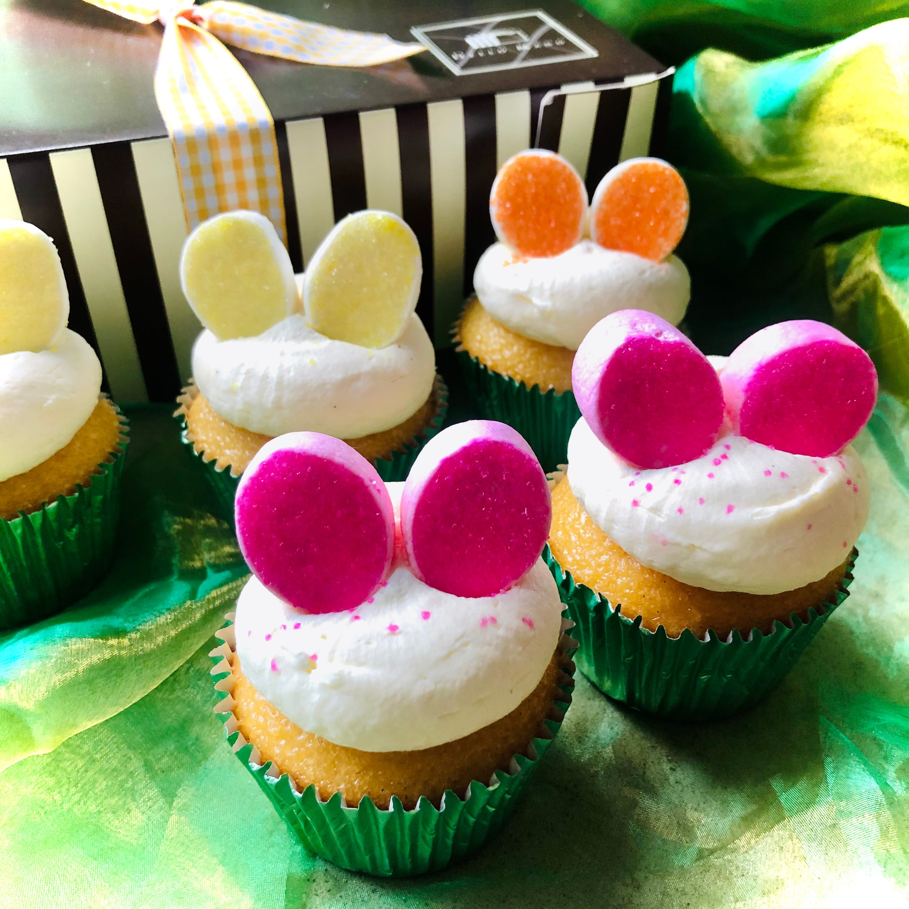 Coloured Easter Bunny Cupcakes with marshmallow ears coloured with sugar in front of a gift box.