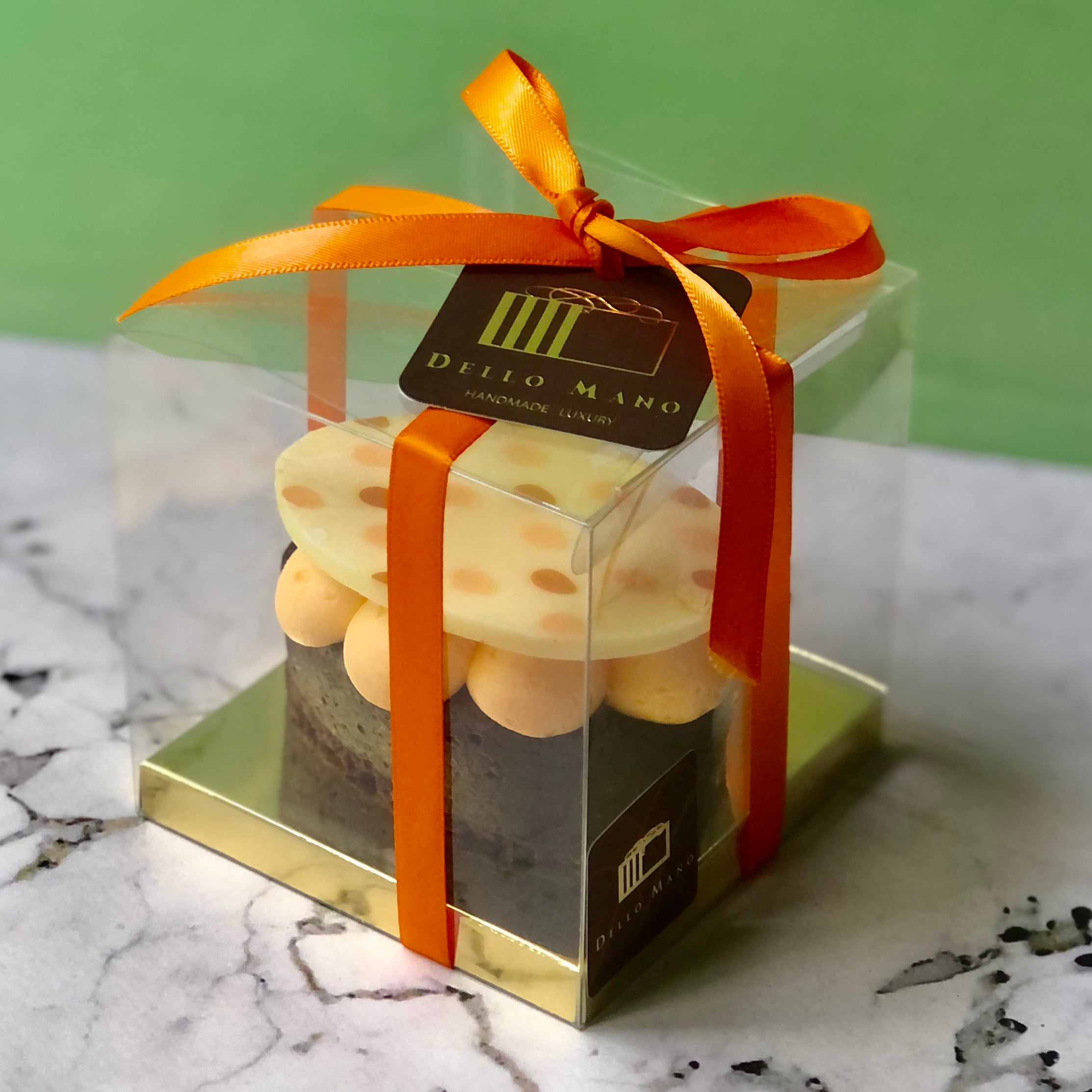 An Easter Brownie Gift Box showing gold base, Easter egg shaped brownie topped with mounds of pastel orange butter cream and an orange spotted white chocolate top.of 