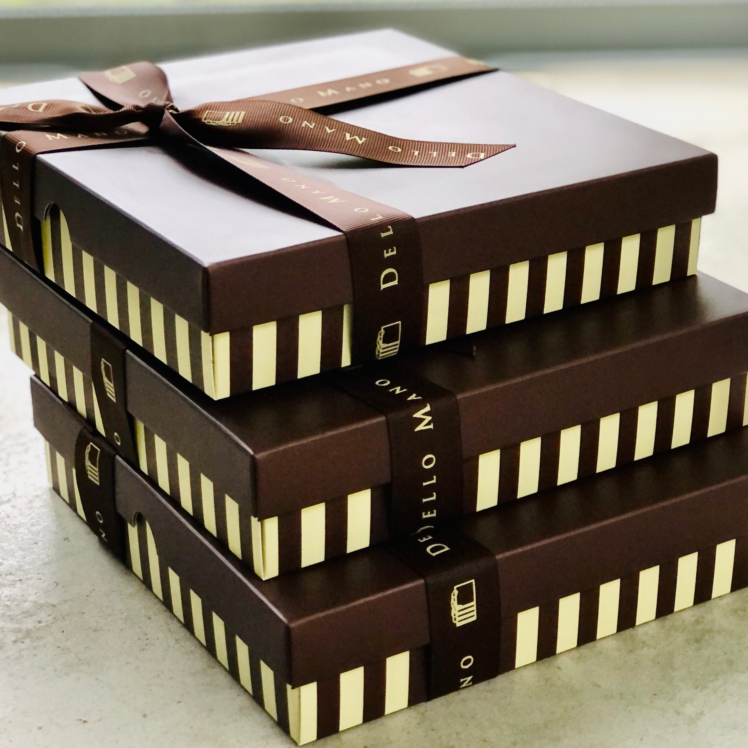 A stack of 3 stripped chocolate brownie gift boxes each with grossgrain ribbon that is embroidered to say Dello Mano