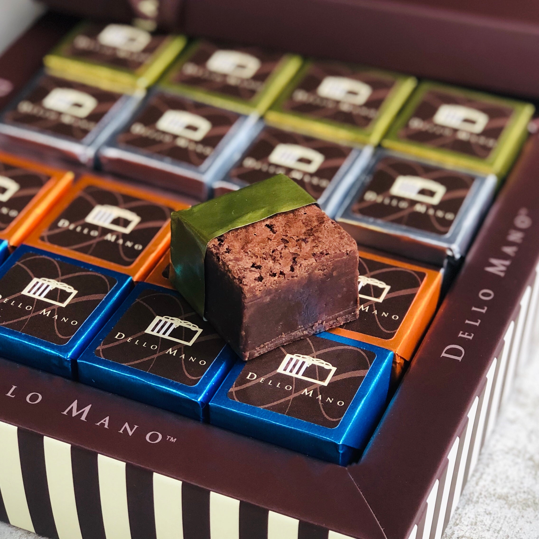 An open chocolate gift box of colourful foile wrapped brownies. A gold foiled brownie is open on top showing a rich brownie cube inside. The striped gift box says Dello Mano