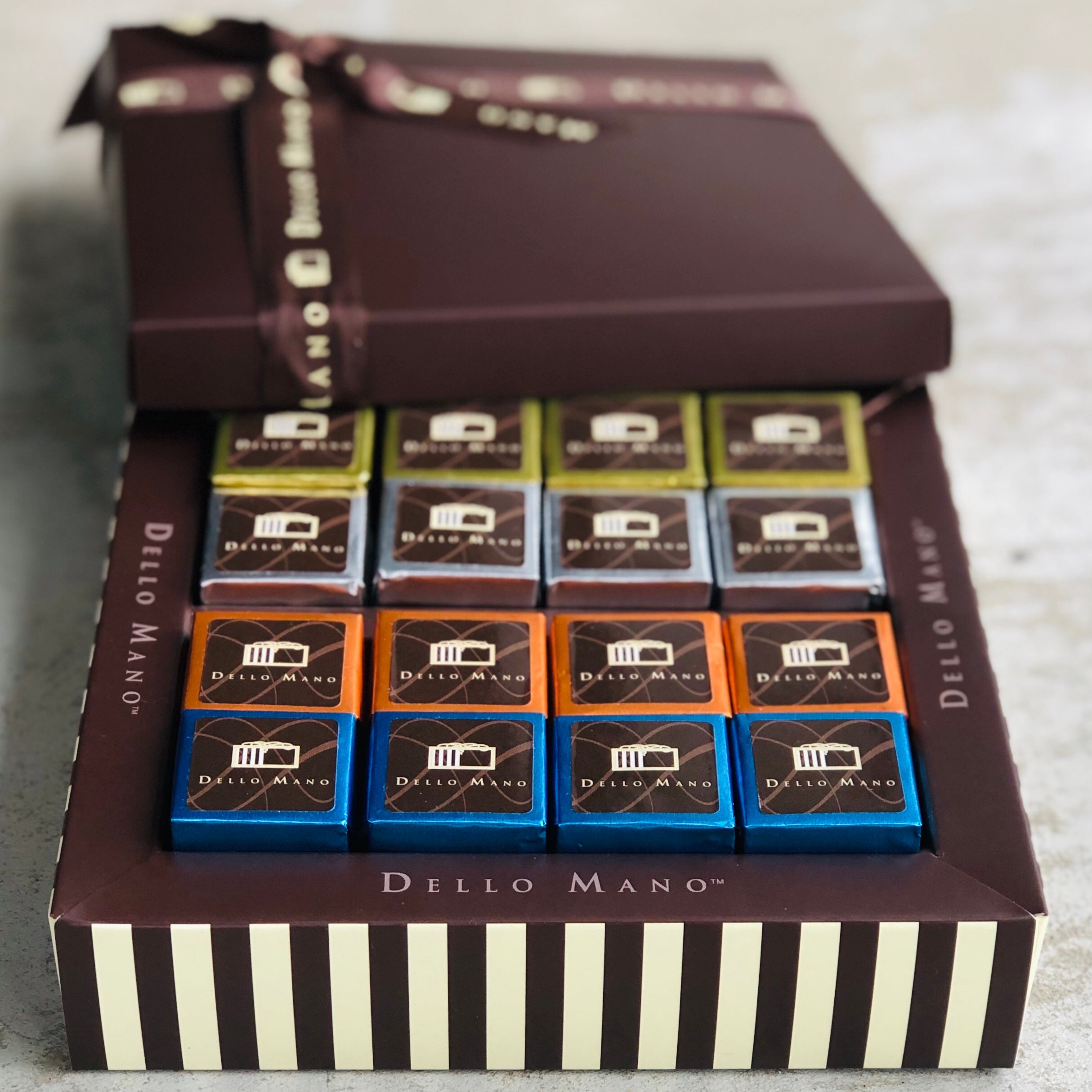 Four rows of four colourful foiled brownies in a chocolate gift box. Rows of blue, orange, silver and gold foiled brownies are tucked inside the striped chocolate gift box. The box has words saying Dello Mano