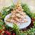 A gingerbread christmas tree with white icing and raspberry/gold sprinkles on a white plate surrounded by greenery