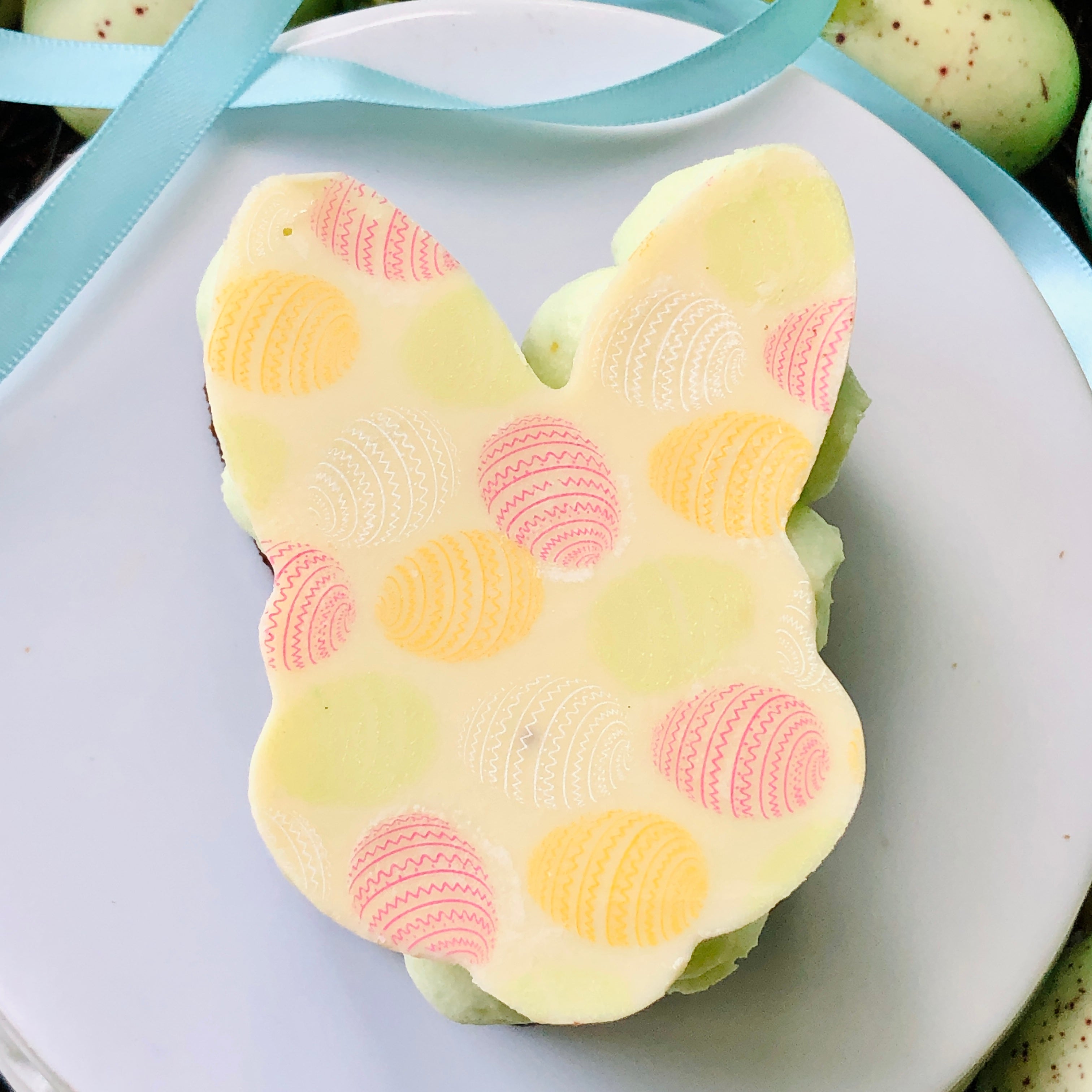 The top view of a Easter Bunny Brownie gift. Sitting on a white plate the top of the Easter gift can be seen. The top is a white chocolate Easter bunny shape with Easter egg patter on top. There are Easter Eggs in the background