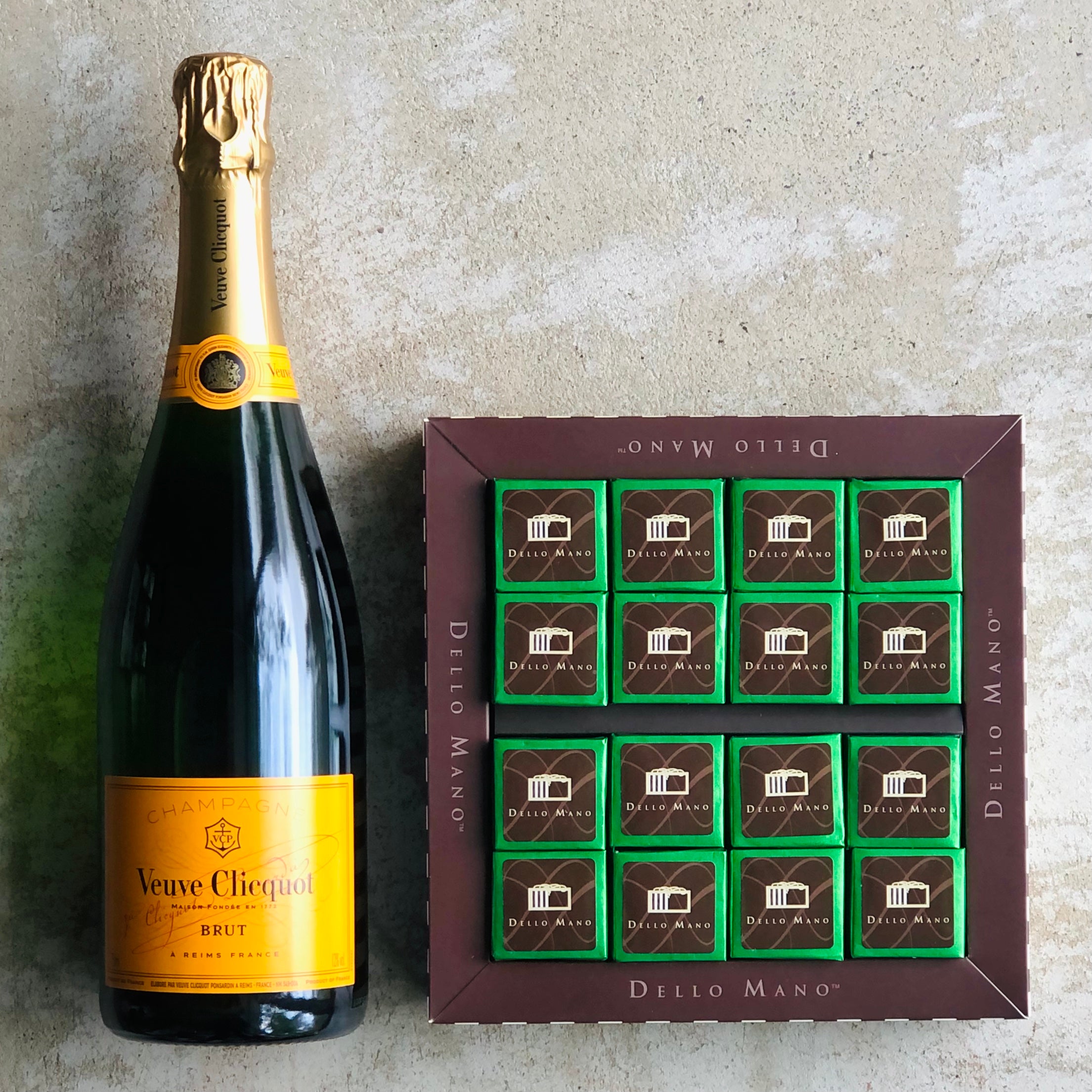 A chmpagne bottle sitting beside an open chocolate box filled with green foiled individually wrapped vegan brownies. The brown chocolate box has the words Dello Mano printed on it.