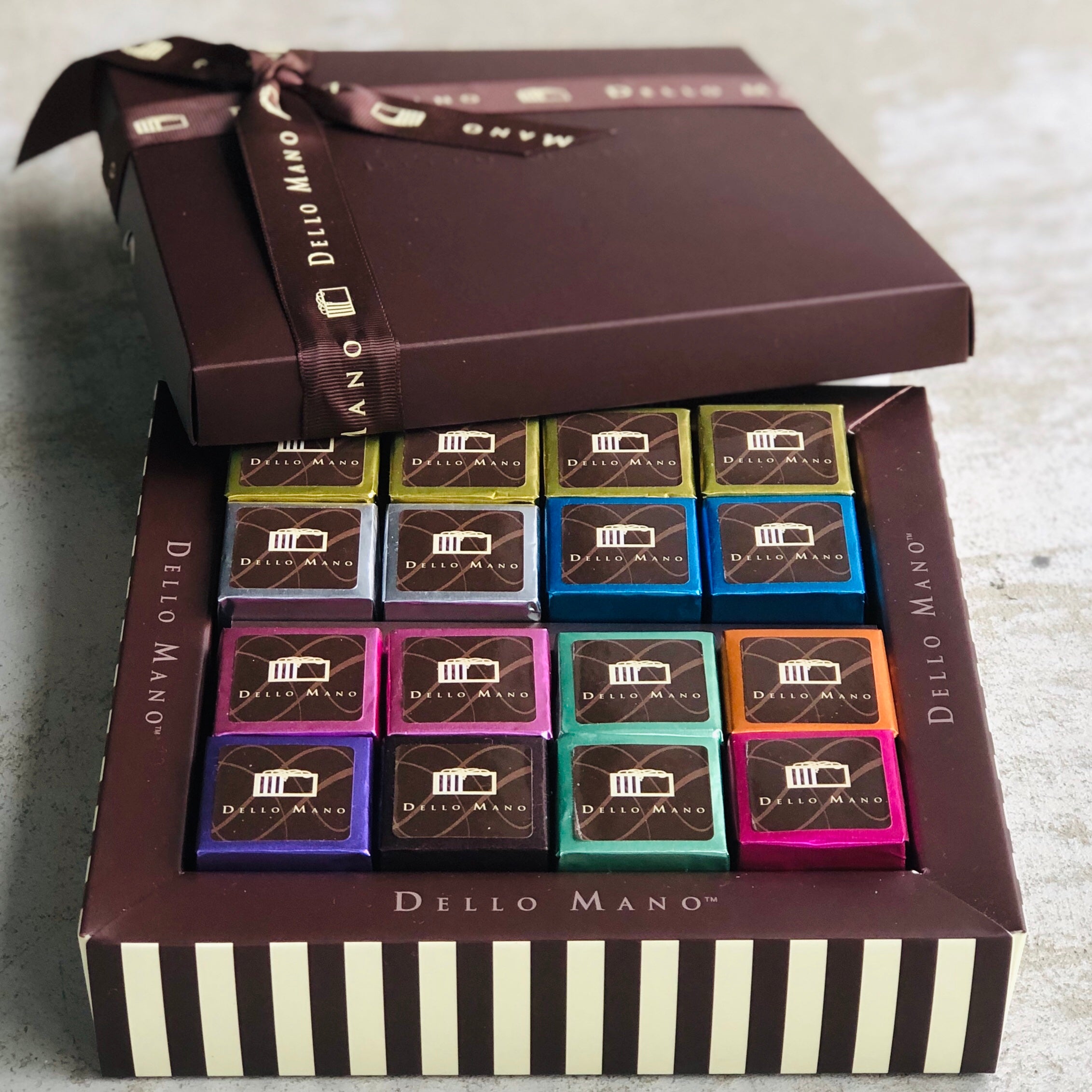A mixed box of brownies with colourful foils. The chocolate gift box has the lid open and a ribbon that has words saying Dello Mano