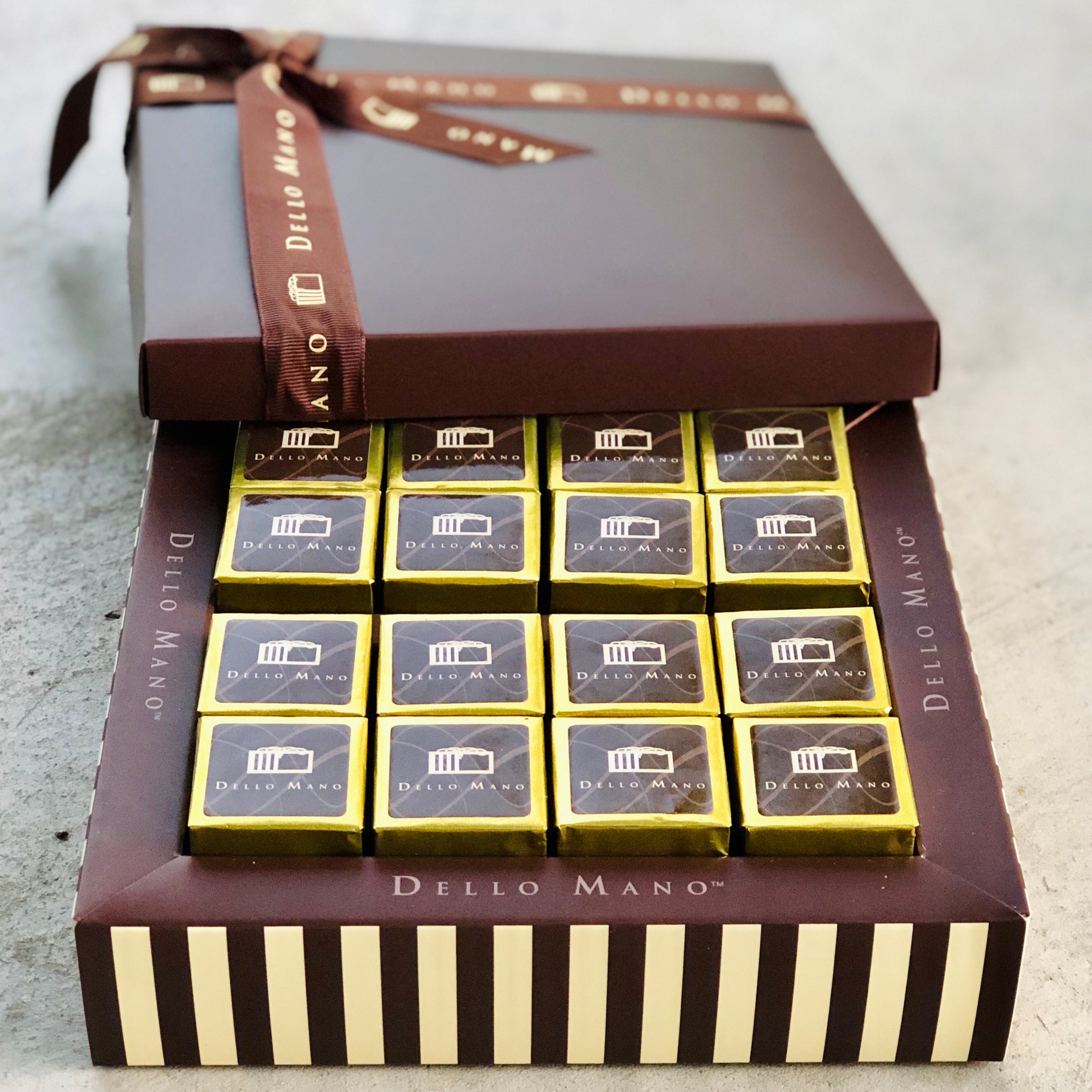 16 pieces of gold foiled Classic Brownie in a  beautiful striped Chocolate gift box. The box has Dello Mano written on it. The lid is off the box and is tied with brownie ribbon