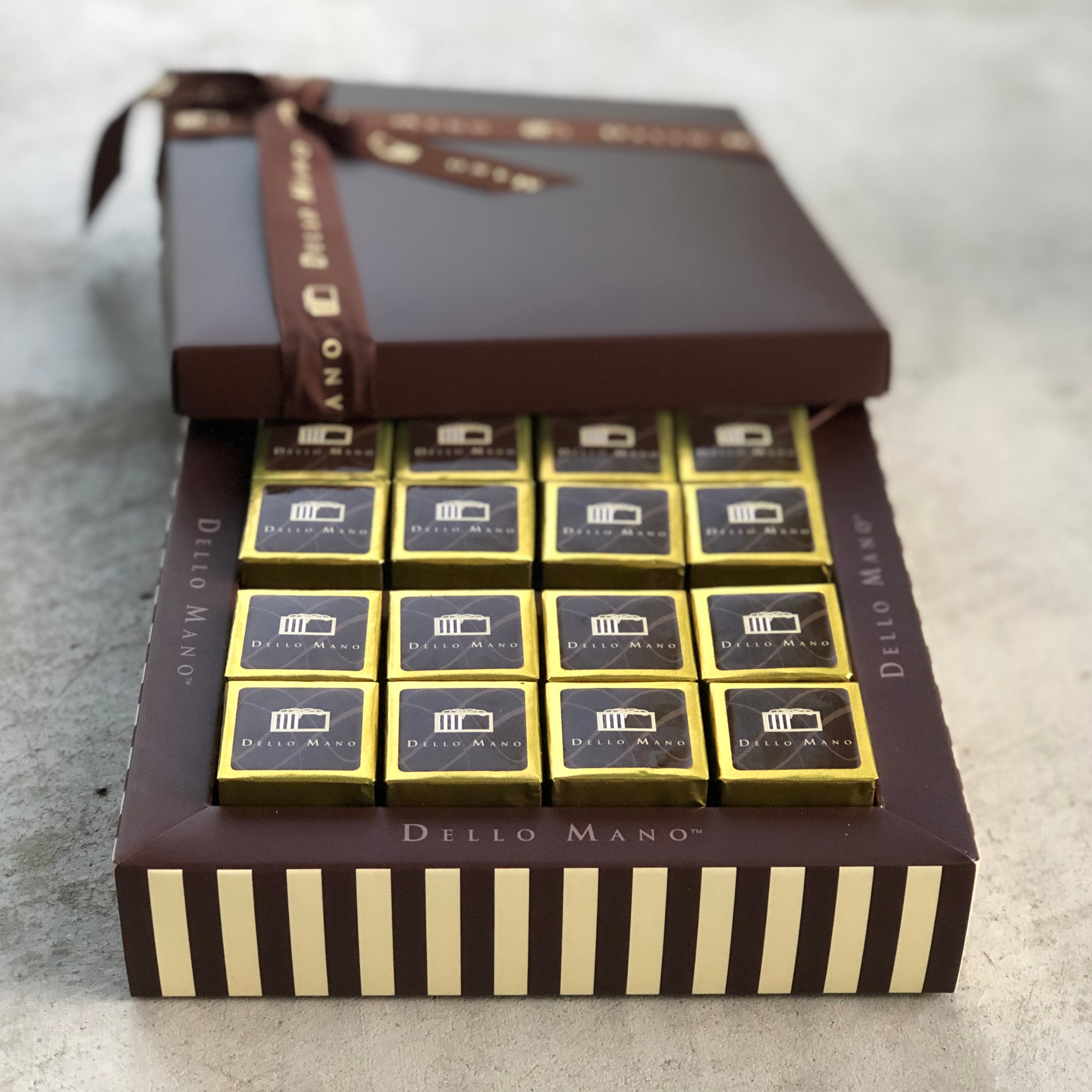 An open Brownie Chocolate gift box showing gold individually wrapped Brownie cubes. The side of the box is striped and sits on a concrete counter top
