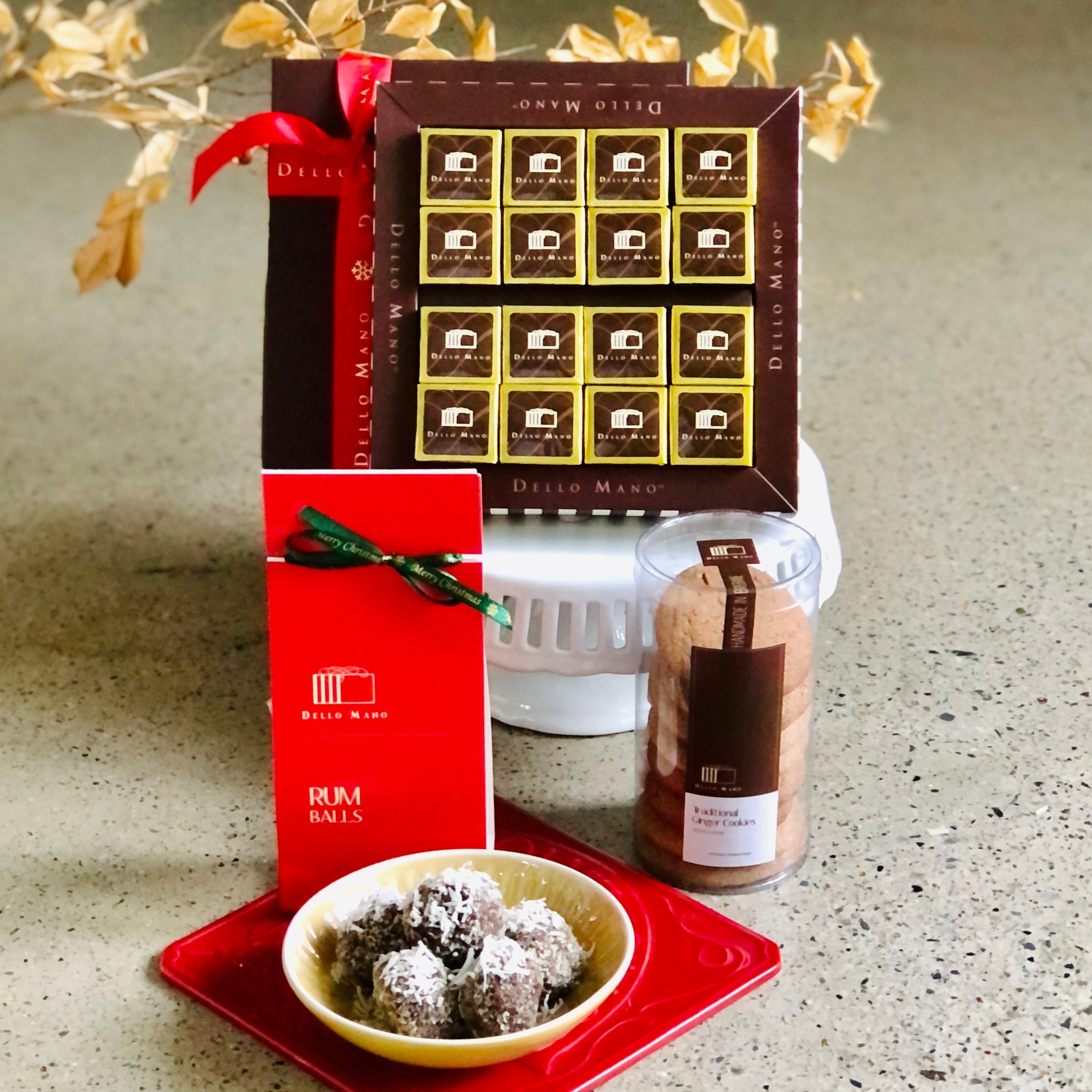 A white cake stand with a box of gold foiled brownies in a gift box. The box says Dello Mano. In front is a pack of rum balls and tube of ginger cookies. In front again is an open bowl of rum balls on a red plate. 