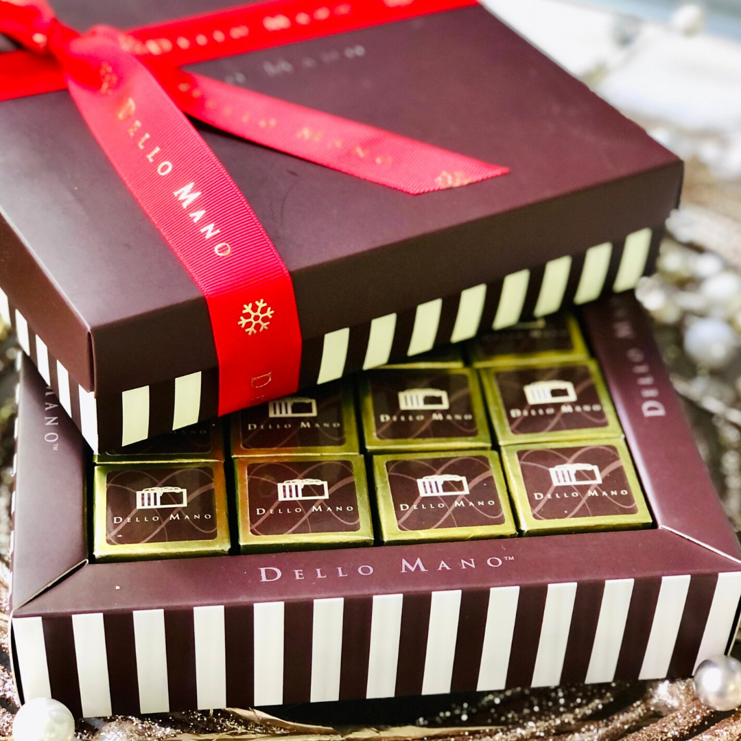 A chocolate gift box with gold foiled brownies that say Dello Mano on the label.  Another box sits on top, this one closed with red ribbon that also says Dello Mano. the ribbon has a Christmas motif.
