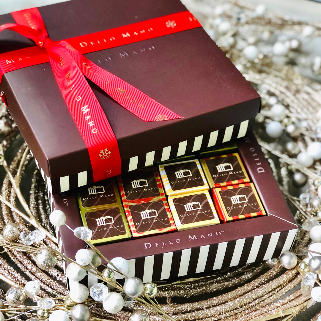A chocolate gift box with stripes filled with gold coloured foiled brownies. On top is a lid also striped with red ribbon, Christmas snowflake motif and the words Dello Mano printed on it.