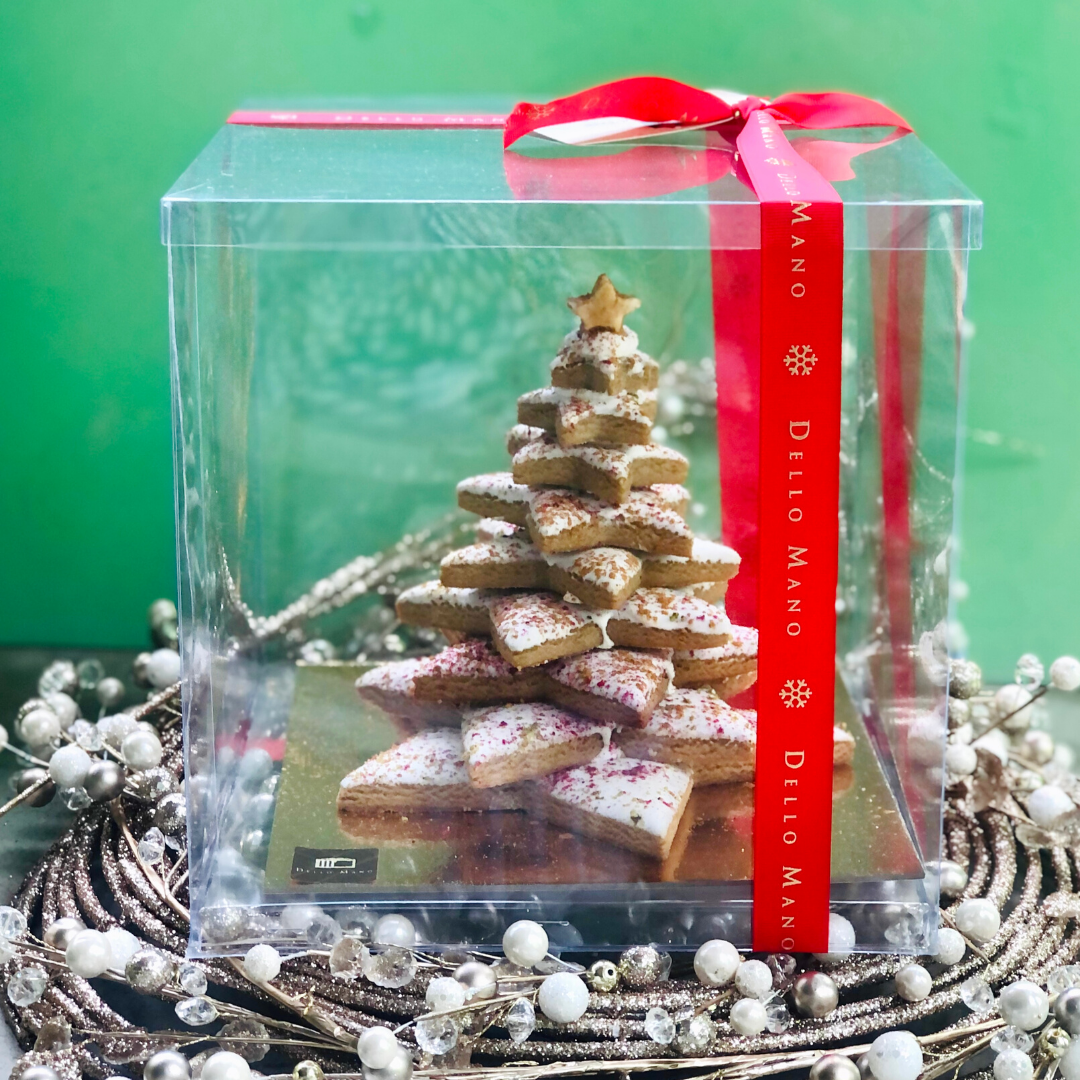 A gingerbread Christmas tree in a clear presentation gift box with red ribbon that says Dello Mano. On a gold and white Christmas wreath