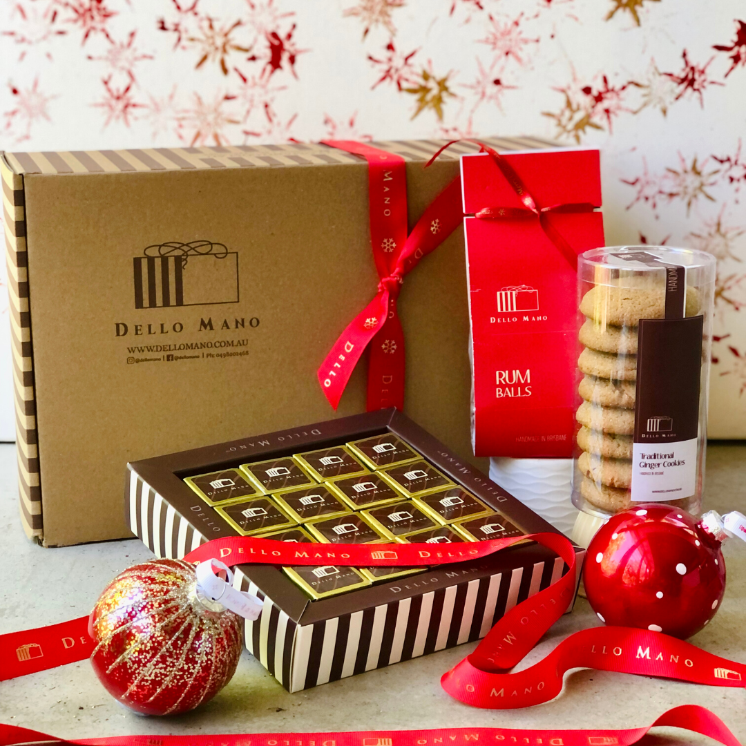 A hamper box with contents layed in front. A box of gold foiled brownies nestled in a striped chocolate box, red ribbon drapes across it. The ribbon says Dello Mano. Also there is a packet of Rum Balls, a canister of Gingerbread cookies and 2 Christmas baubles.