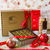 A hamper box with the words printed Dello Mano. In front a gift box of gold foiled brownies, a red bowl of rum balls and a pack of ginger cookies. Two Christmas baubles sit in the forefront of the image. 